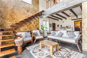 Un mur en pierre, un escalier en bois, des chaises en bois, un canapé, une table en bois et un tapis à motifs dans une piÚce.