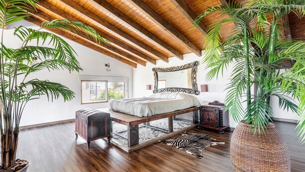 A bedroom with a wooden ceiling, a bed with a white mattress, a large mirror, and a potted plant.