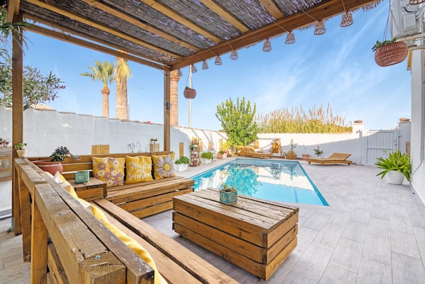 An outdoor pool area with wooden furniture, a pergola, and potted plants.