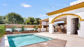 Un espace piscine doté d’une terrasse en bois, d’un coin salon abrité et d’un bâtiment surmonté d’un toit en tuiles.