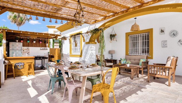 Un espace extérieur abrité, aménagé pour les repas, avec une table et des chaises en bois, un coin bar et un espace détente avec un canapé et des fauteuils.