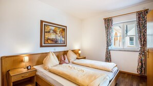 Un lit double avec une tête de lit en bois, une table de chevet avec une lampe, une commode, un tableau au mur et une fenêtre avec des rideaux.