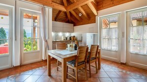 Una cocina con vigas de madera, una mesa de comedor con sillas y ventanas grandes con cortinas.