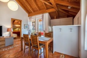 Ein Essbereich mit Holzkstühlen und einem Tisch, eine Küche mit weißen Schränken und eine Decke mit freiliegenden Balken.
