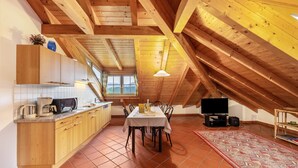 Habitación estilo ático con techos altos, vigas de madera, una mesa de comedor, sillas, televisor y una zona de cocina con armarios y electrodomésticos.