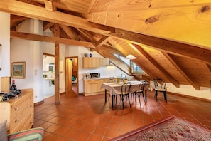 An attic room with exposed wooden beams, a dining table with chairs, a kitchen area, and a view through a window.