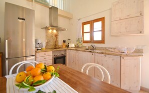 A kitchen with a stainless steel refrigerator, a stove, a sink, and a window with curtains. There is a table with a bowl of oranges.