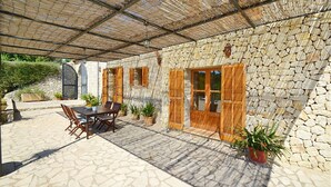 A stone building with wooden shutters, a covered patio with a table and chairs, and potted plants. - Country House 'Sa Volta' with Mountain View, Wi-Fi and Air Conditioning (selva)