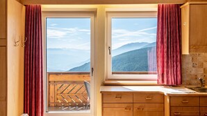 A kitchen with wooden cabinets, a window with a view of mountains, and a balcony with a wooden railing. - Studio Apartment 'Spinges' with Mountain View, Private Terrace and Wi-Fi (Maranza)