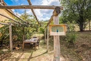 A wooden pergola structure with a table and chairs underneath, surrounded by greenery and a clear sky.