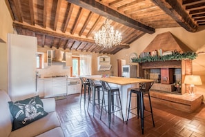 A kitchen with a wooden ceiling, a central island with bar stools, a fireplace, and a bookshelf.