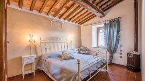 A bedroom with a wooden ceiling, a bed with a headboard, bedside tables, a window with curtains, and a vintage radio. - Holiday Apartment 'Glicine' with Mountain View, Shared Pool & Wi-Fi (Montieri)
