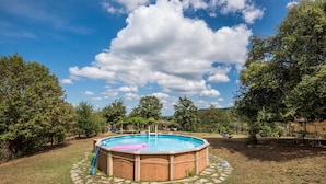 Una piscina circular con muelle de madera y escalera, rodeada por un camino de piedra y árboles.