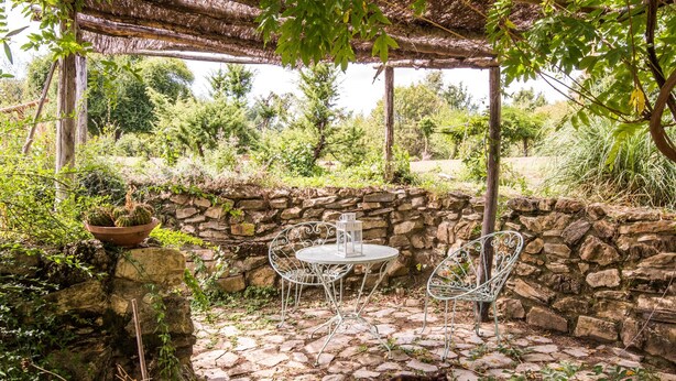 Un patio de piedra con sillas y una mesa de metal, rodeado de follaje y un muro de piedra.