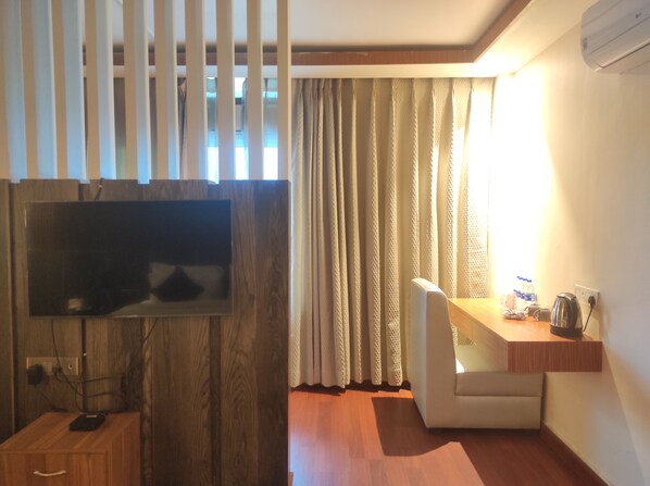 A room with a flat-screen TV mounted on a wooden panel, a small desk with a kettle, and a sofa.