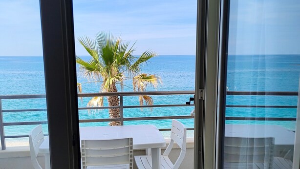 Un balcón con mesa y sillas, con vistas a una playa de palmeras y al mar.