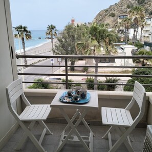 Un balcon avec deux chaises blanches et une table, donnant sur une plage et une colline rocheuse.