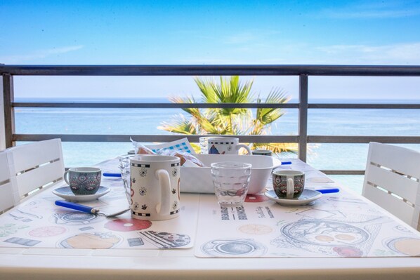 Une table sur laquelle sont disposés divers mugs et tasses, un verre et une cuillère, le tout sur fond de ciel bleu dégagé et de vue sur l’océan.
