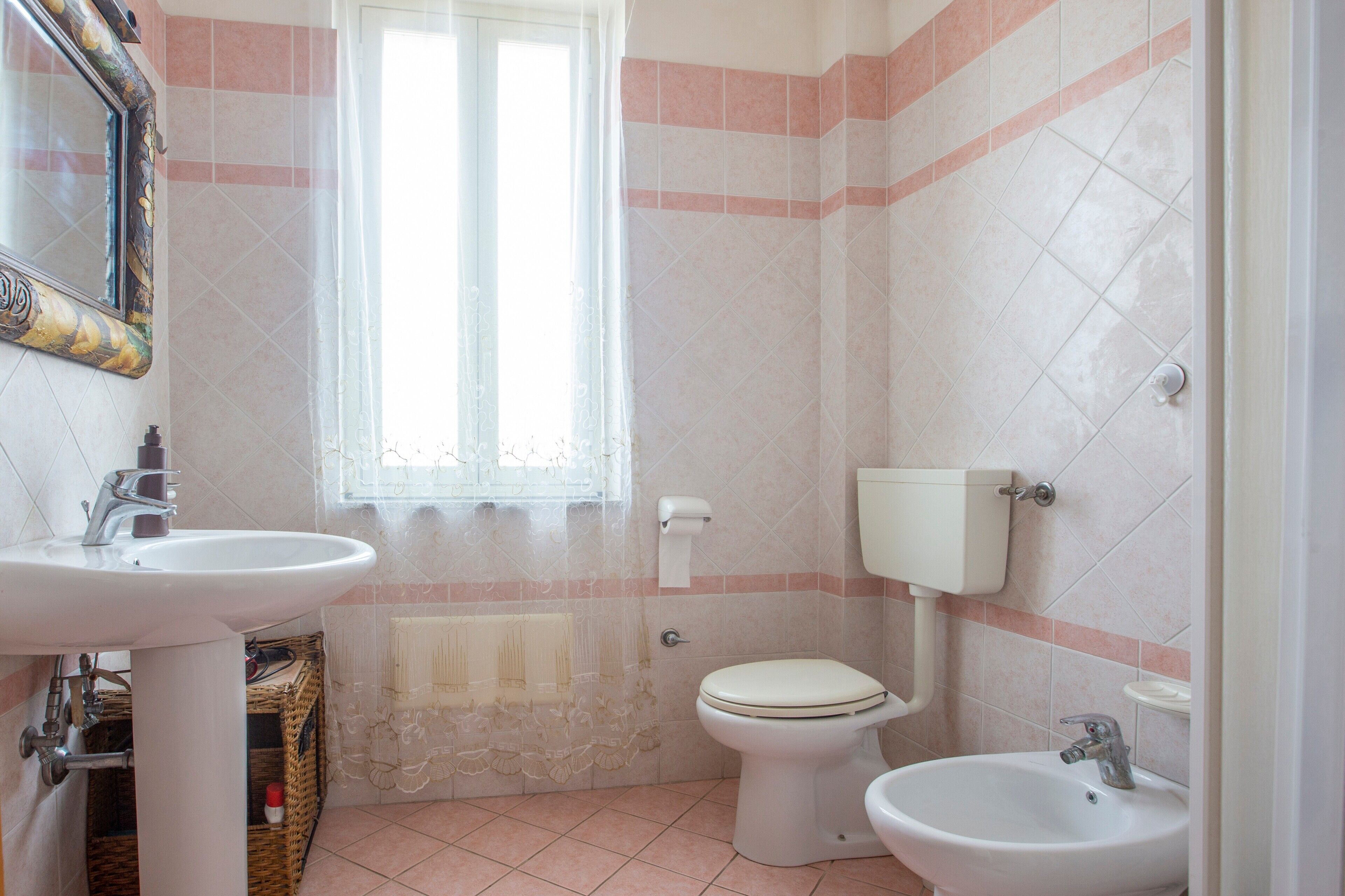 Een badkamer met een wastafel, toilet en bidet, een raam met kantgordijnen, en een betegelde vloer.