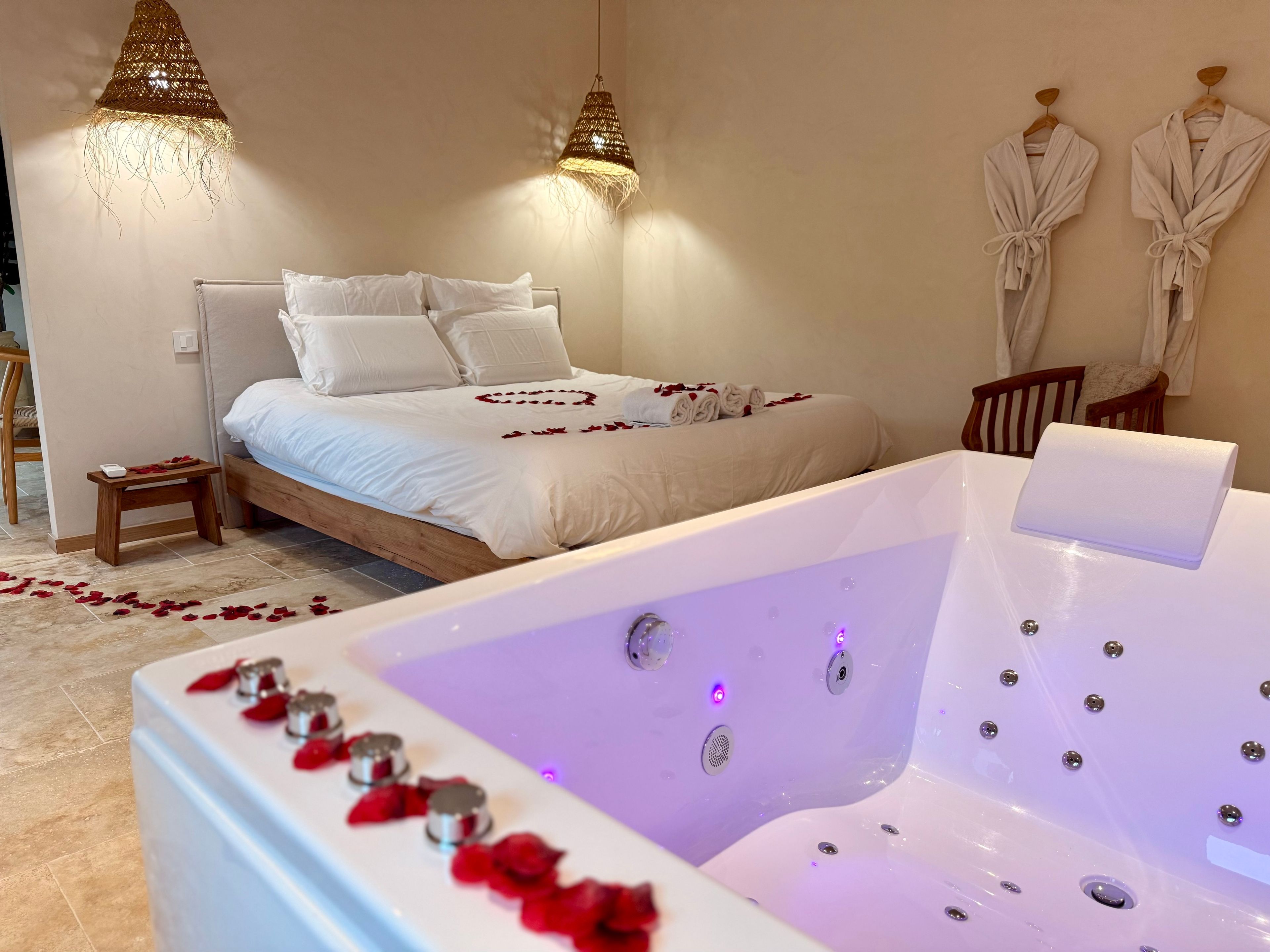 Un dormitorio con cama, mesita de noche, una bañera grande con pétalos de rosa y dos lámparas de pared.