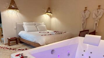 Un dormitorio con cama, mesita de noche, una bañera grande con pétalos de rosa y dos lámparas de pared.