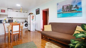 Una sala de estar con un sofá marrón, una mesa de comedor con sillas, una cocina con armarios blancos, un refrigerador y un cuadro de una playa.