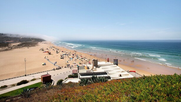 Una playa con vista clara al océano, una orilla arenosa y algunos edificios al fondo.