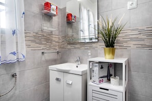 Ein Badezimmer mit einem weißen Waschbecken, Spiegel und Dusche. Im Waschtischbereich befinden sich Ablageflächen und ein Blumentopf.