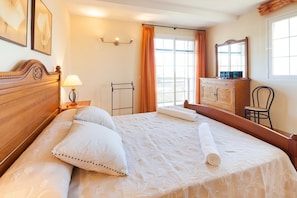 Una camera d'albergo con un letto, un comodino con una lampada, un settimino in legno, una sedia e una finestra con tende.