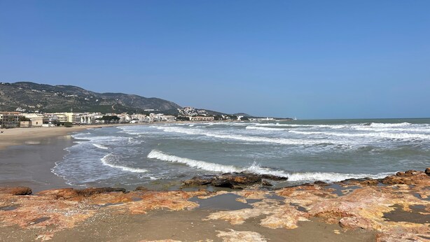 Une plage avec des vagues, une côte rocheuse et des bâtiments en arrière-plan.