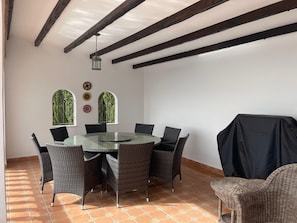 Spiseområde med et rundt glasbord omgivet af fletstole, et loft med synlige bjælker og et vindue med udsigt til grønne omgivelser.