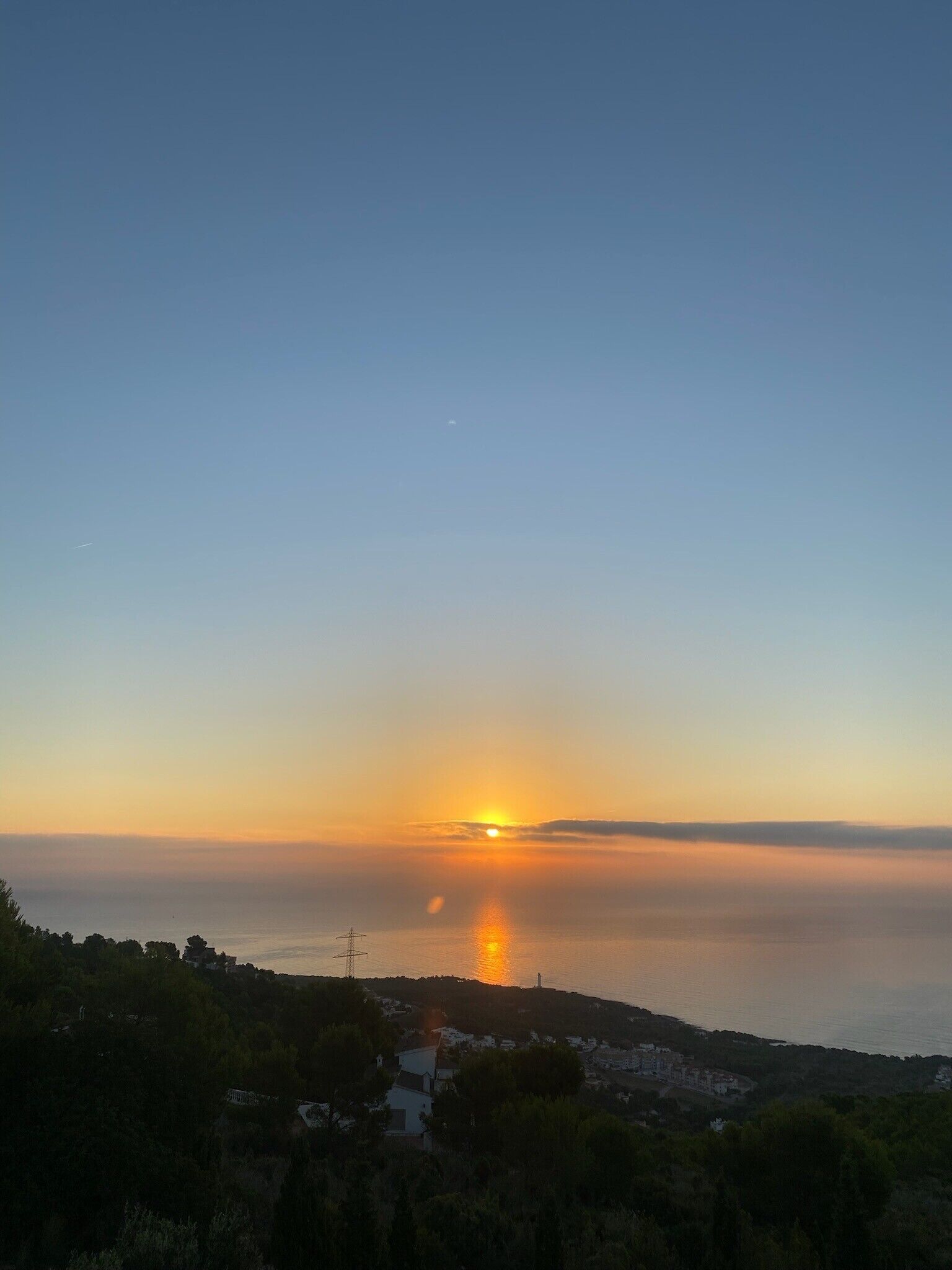 Een uitzicht op een kustgezicht bij zonsondergang, met een heldere lucht, een kalme zee en een verre elektriciteitsmastenstructuur.
