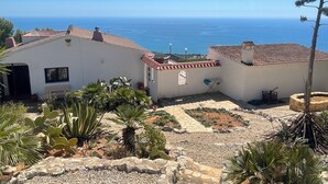 Un bâtiment blanc surmonté d’un toit de tuiles rouges, un petit jardin agrémenté de cactus et de plantes tropicales, et une vue sur la mer.