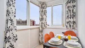 Un comedor con una mesa preparada para dos personas, dos sillas anaranjadas, un ventanal con vistas a edificios y un radiador debajo de la ventana.