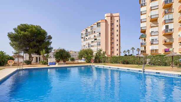 Una piscina con agua cristalina azul, rodeada de un jardín cuidado y un edificio residencial alto al fondo.