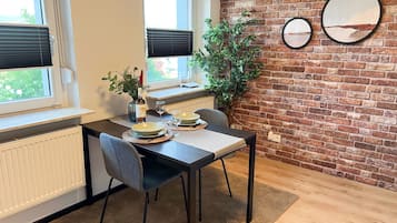 Een eethoek met een bakstenen muur, twee ramen met zonwering, een potplant en een gedekte tafel voor twee personen.