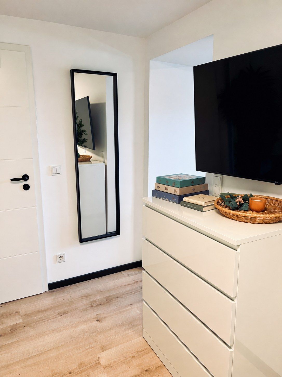 A room with a white wardrobe, a flat-screen TV mounted on the wall, and a mirror reflecting the TV.