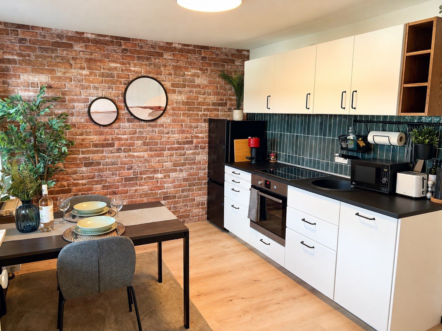 Cocina moderna con armarios blancos, pared de ladrillo, mesa de comedor con sillas y una planta pequeña.