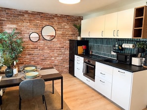 A modern kitchen with white cabinets, a brick wall, a dining table with chairs, and a small plant. - JUPAS° Studio21 – Terrace with Hanging Nest (Trier)