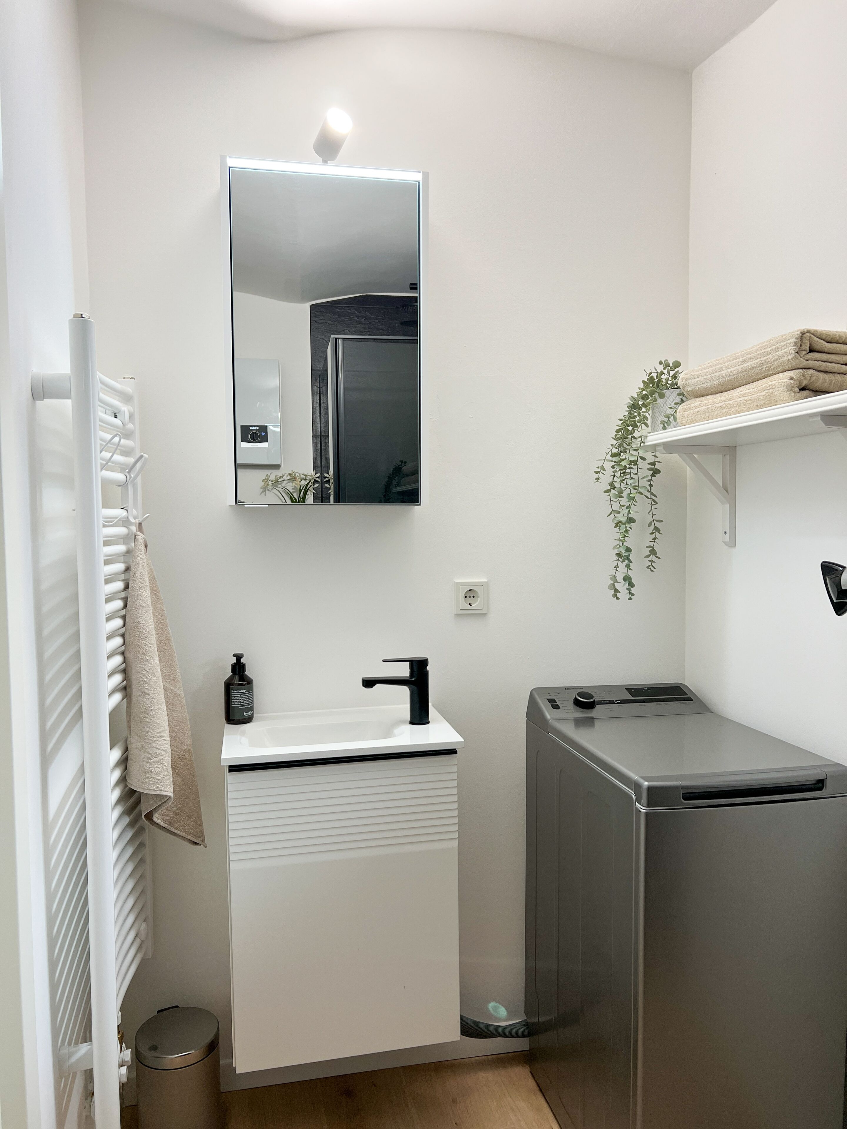 Un baño moderno con tocador blanco, espejo, perchero, lavamanos, inodoro y un pequeño cubo de basura.