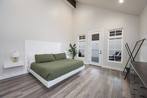 A bedroom with a bed, a nightstand, a lamp, a plant, and a view of the outside through a door.