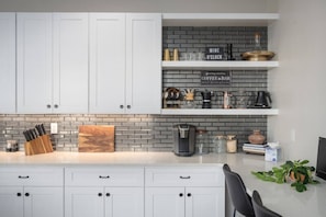 A modern kitchen with white cabinets, a gray brick backsplash, and a coffee bar area with various coffee-making equipment.