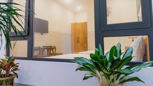 Une pièce comprenant un lit, une armoire en bois, une table et des chaises. On y trouve également des plantes en pot et une fenêtre donnant sur une autre pièce.