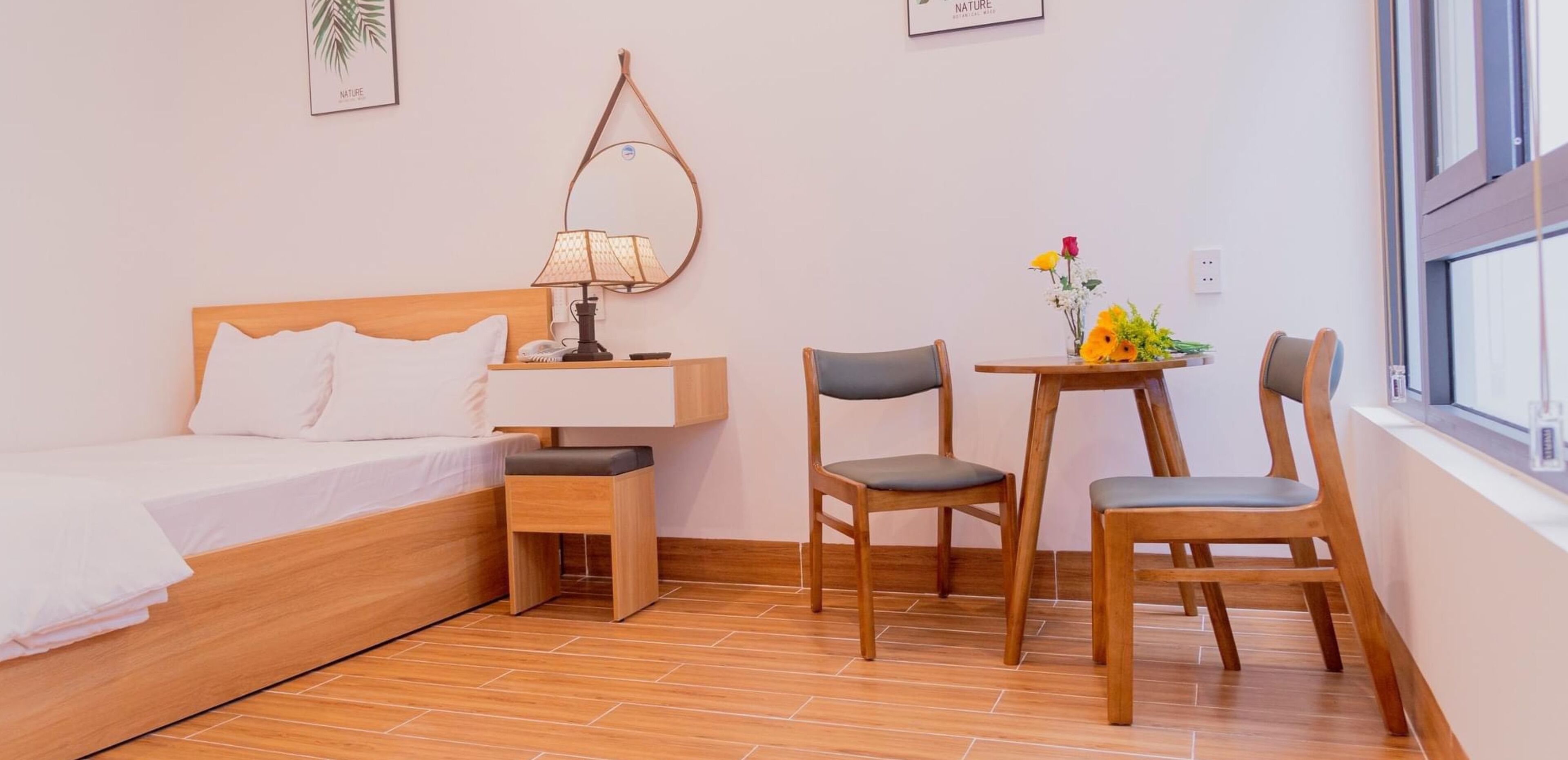 A hotel room with a bed, a bedside table with a lamp, a small table with chairs, and a window with a view.