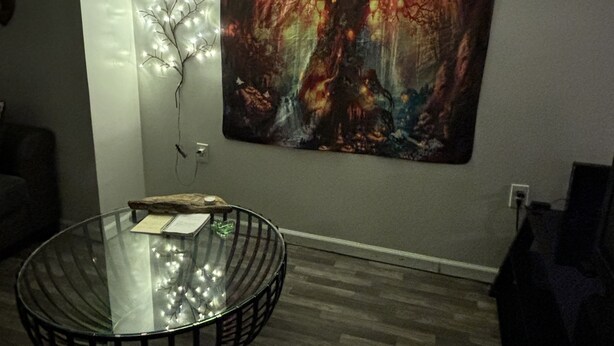 Um quarto com uma mesa de vidro redonda, um tapeçário com uma cena de floresta e um respiradouro no teto.