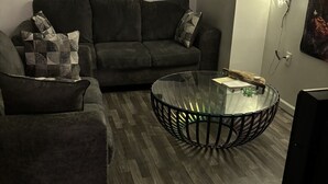 Uma sala de estar com um sofá escuro, uma mesa de centro de vidro e uma televisão de tela plana.