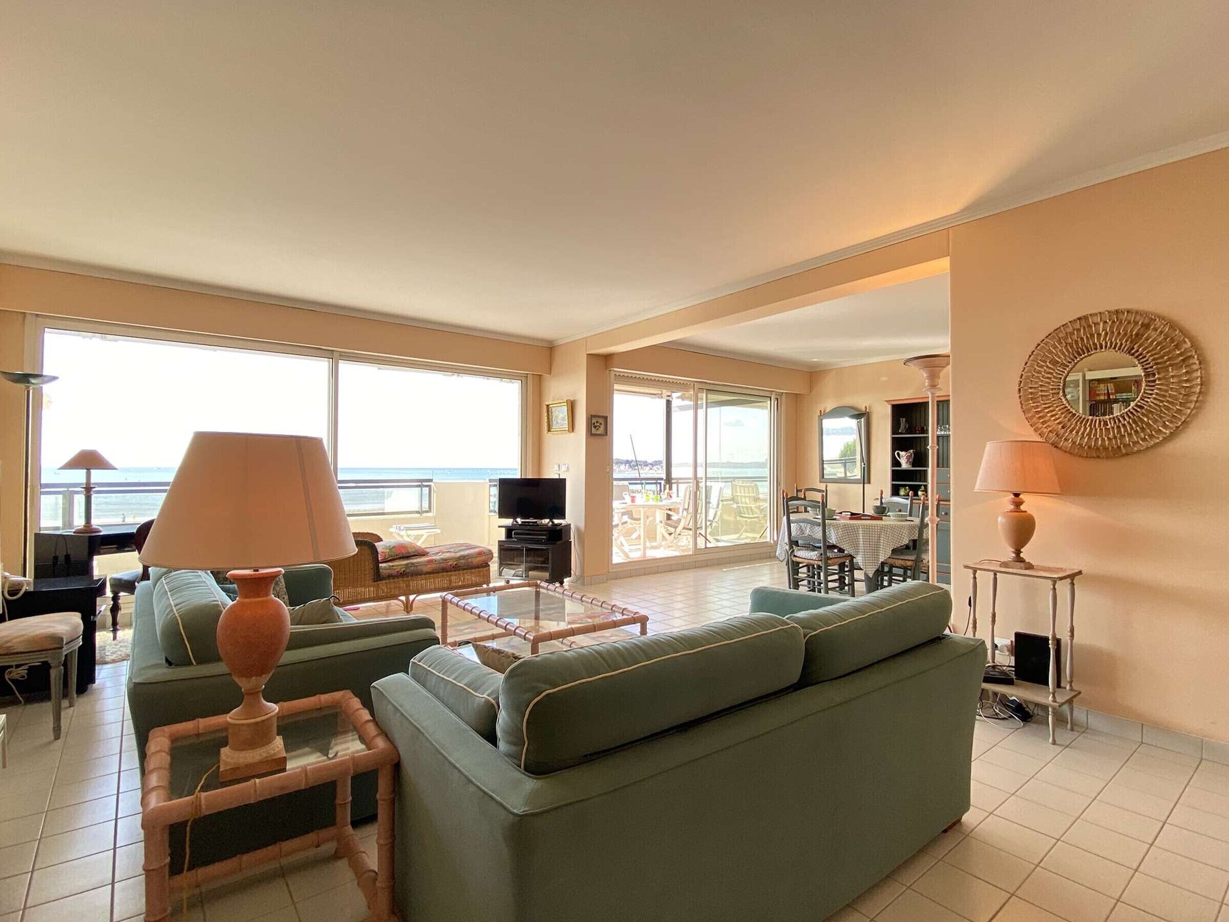 A spacious living room with a large sofa, a coffee table, and a television. There is a balcony with a view of the sea.
