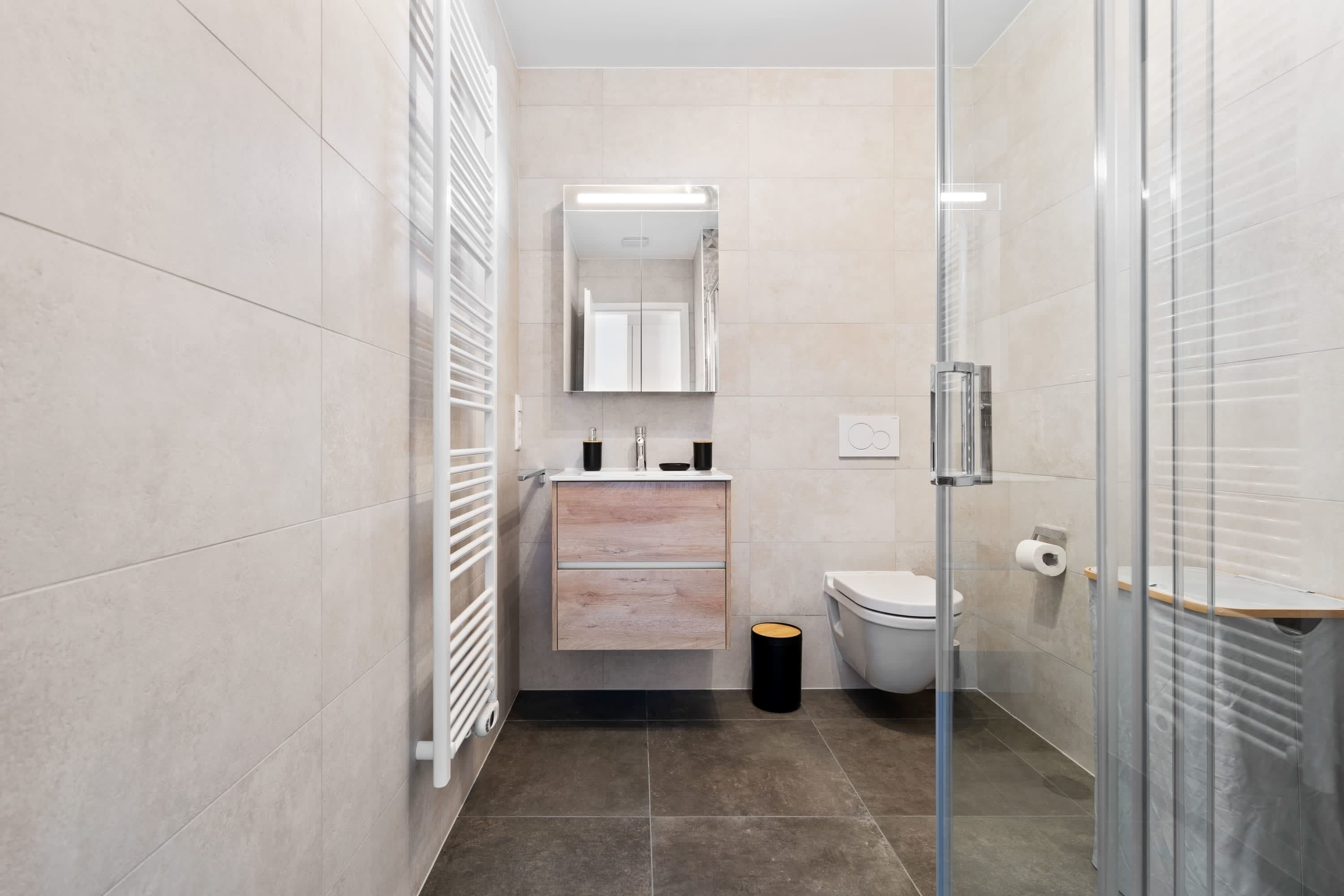 A modern bathroom with a glass shower enclosure, a white toilet, a vanity with a mirror, and a black trash bin.