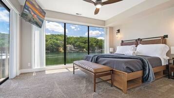 A bedroom with a large window, a ceiling fan, a bed, a bench, and a view of a lake.