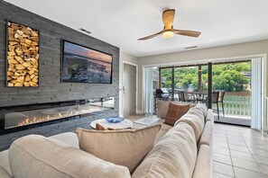 A modern living room with a fireplace, a flat-screen TV, a ceiling fan, and a view of a balcony. - Lake Life Location with Serenity (Osage Beach)